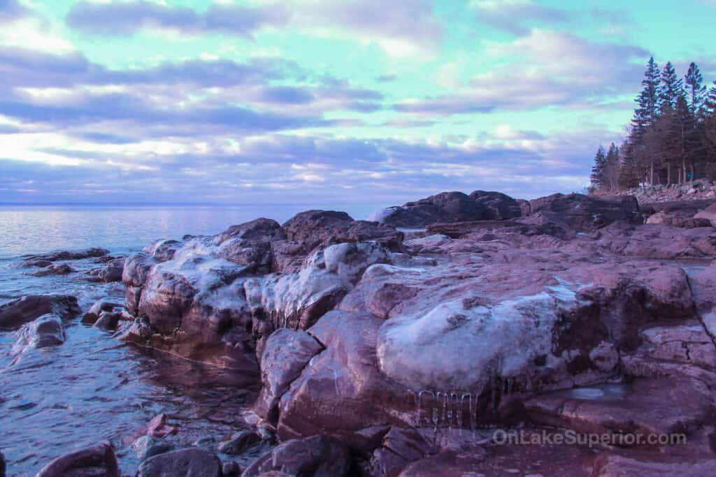 photo with icy rocks in the foreground with the waters of Lake Superior leading to the cloud-masked sunrise in the background
