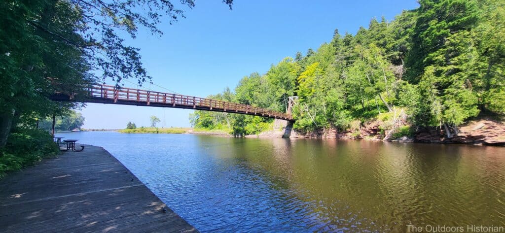 A view of the suspension bridge from the path on the west side of the river on a sunny day.